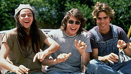 Three young adults sitting outdoors, smiling and gesturing, two wearing casual clothes and one in overalls, trees in background