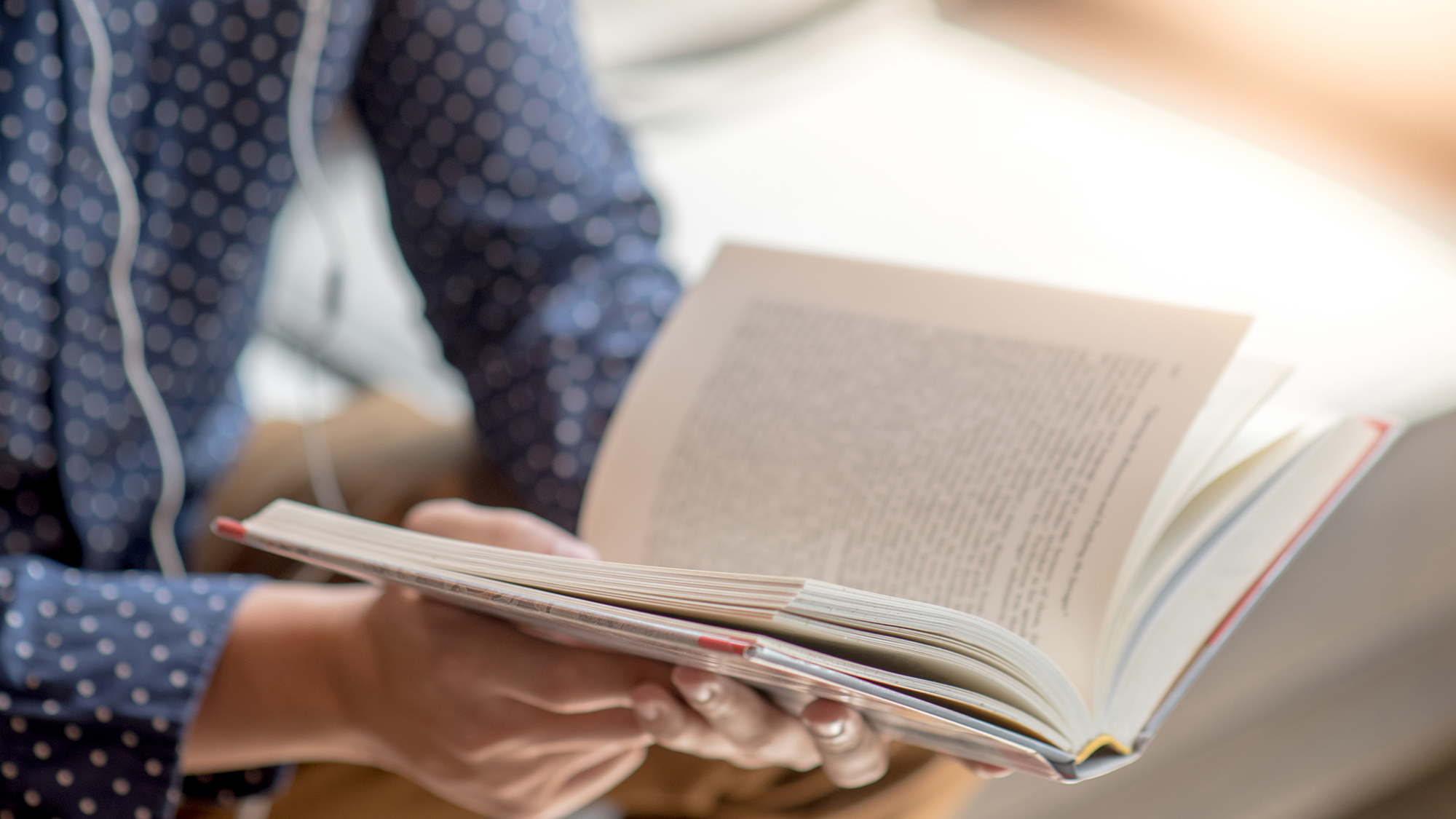 Person in a polka dot shirt reading an open book near a bright window